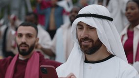 Handsome arabian man having video call on smartphone while watching football game on stadium. Dedicated fan sharing with feelings and emotions, during online communication.