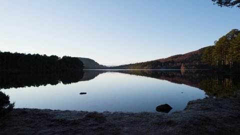 Aerial drone footage rising over the surface of still water (Loch an Eilein) with reflections in the Cairngorms National Park, Scotland to reveal a native forest and mountain landscape at sunset
