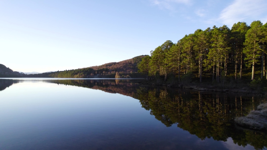 Aerial drone footage flying over the surface of still water (Loch an Eilein) with reflections in the Cairngorms National Park Scotland towards a native scots pine forest with clear blue sky at sunset. Royalty-Free Stock Footage #1085310587