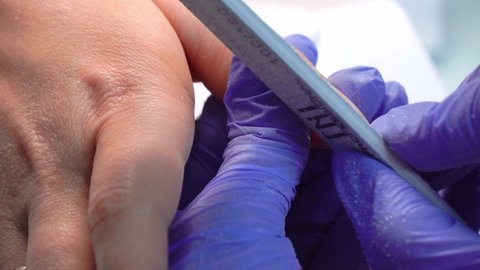 Close-up of a manicure master in blue gloves filing nails with a special nail file. Nail treatment process.