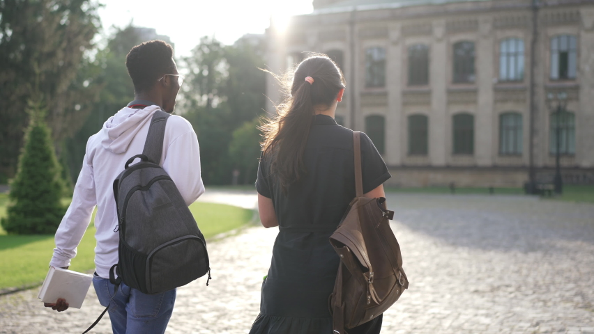 Back view young interracial couple of students walking on sunny morning to university campus outdoors. Confident smart African American man and Caucasian woman strolling talking in sunshine | Shutterstock HD Video #1085313446