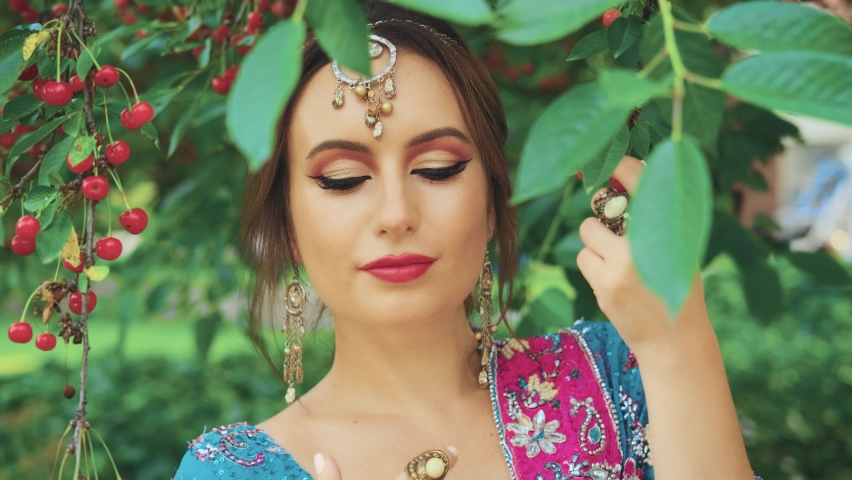 Closeup portrait oriental fantasy woman. India princess gently touches lips cherry red berry. Traditional asia pink makeup. National vintage blue dress. Backdrop summer nature garden green tree leaves