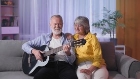 musical hobby, joyful elderly couple play guitar and sing, loving mature woman kisses on cheek of her beloved old husband while sitting on couch in evening in room while relaxing