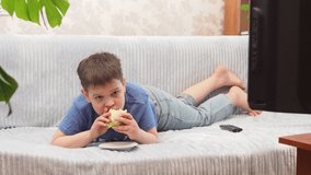 a boy at home on the couch watches TV and eats a sandwich, switches programs with the remote control. Joyful boy playing video games and laughing seated at home.