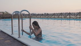 A dark-haired curvy tanned girl with long flowing hair in a bright leopard swimsuit is having fun in a blue pool with clear transparent water, falling into it with her back. 4K UHD slow-mo video
