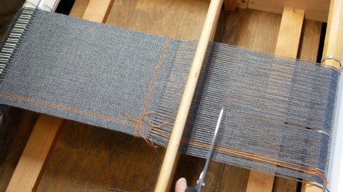 Gray fabric is cutting off of the weaving loom. Hand with scissors cuts the warp threads off the wooden frame. Finishing the woven project. Concept of the hand craft work