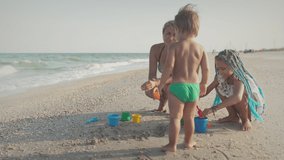 A kind caring beautiful mother plays with her children: eldest skinny daughter and youngest son toddler building beads and sand castles decorating them with shells and pebbles. 4K UHD slow-mo video