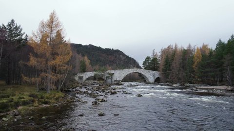 Left rolling aerial drone footage of a fast flowing river (River Dee) and the Bridge of Dee near Braemar and Ballatar in Scotland with Scots pine and European larch trees in autumn on the river banks