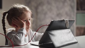 Bored little girl with braided hair watches educative video via tablet on holder sitting at table in light children room closeup slow motion