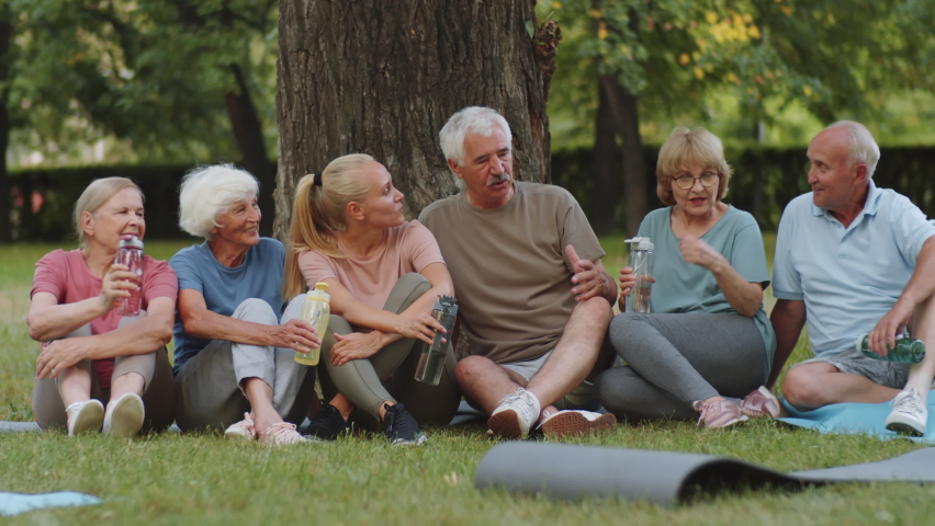 Group of cheerful senior people and young female fitness coach in sportswear sitting together on lawn in park and chatting