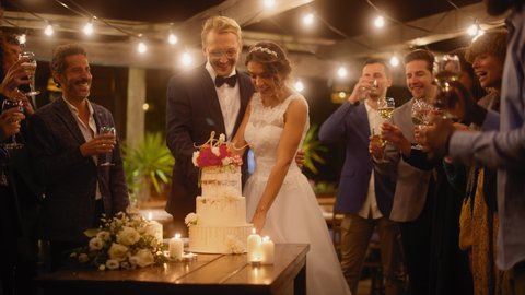 Beautiful Bride and Groom Celebrate Wedding at an Evening Reception Party with Multiethnic Friends. Married Couple Standing at a Dinner Table, Cutting Wedding Cake.