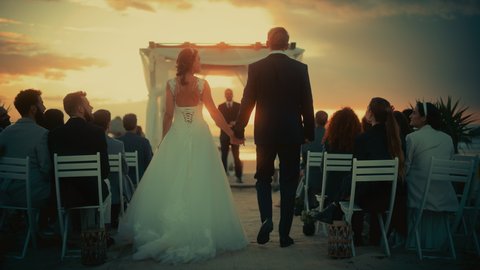 Beautiful Bride in White Wedding Dress and Handsome Groom in Black Suit Going Down the Aisle at an Outdoors Ceremony Venue Near the Sea at Sunset. Happy Multiethnic Friends Celebrating Marriage.