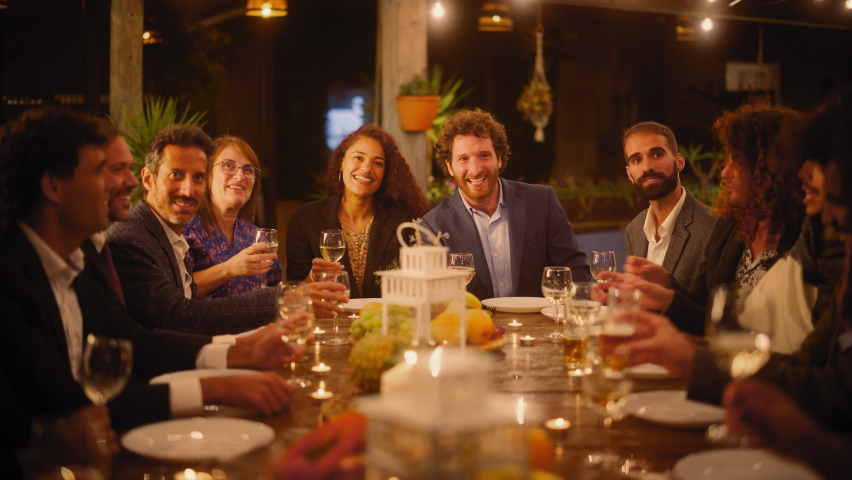 Big Dinner Party with a Small Crowd of Multiethnic Diverse Friends Celebrating at a Restaurant. Beautiful Happy Hosts Propose a Toast and Raise Wine Glasses while Sitting at a Table in the Evening. Royalty-Free Stock Footage #1085341040
