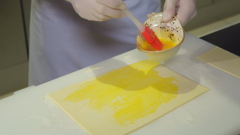 egg yolk covering raw dough layer. fresh dough sheet coating with yellow hen's egg before baking. close-up cook's hands in rubber gloves apply liquid egg on dough by kitchen brush. food preparation