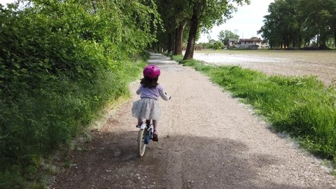 little girl in tulle skirt pink helmet and pink shoes bicicling in country road covered by white tree blossoms from poplar trees in spring season