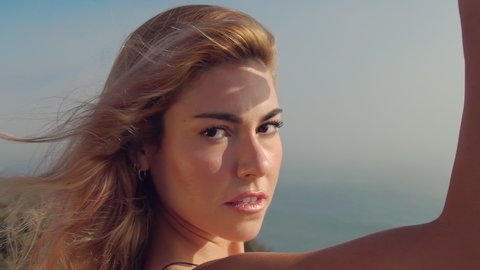 Portrait of a Beautiful attractive blonde posing in slow motion on a rock against the ocean in a black dress. Sexy woman gets pleasure and looks at the camera . The wind blows away the dress