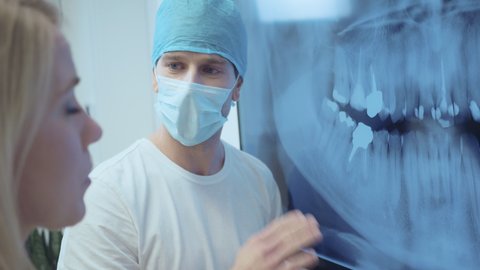 Over the shoulder view of a male dentist talking with female Caucasian patient. The dentist, wearing a protective face mask and surgical cap, explaining the x-ray visible on the screen.