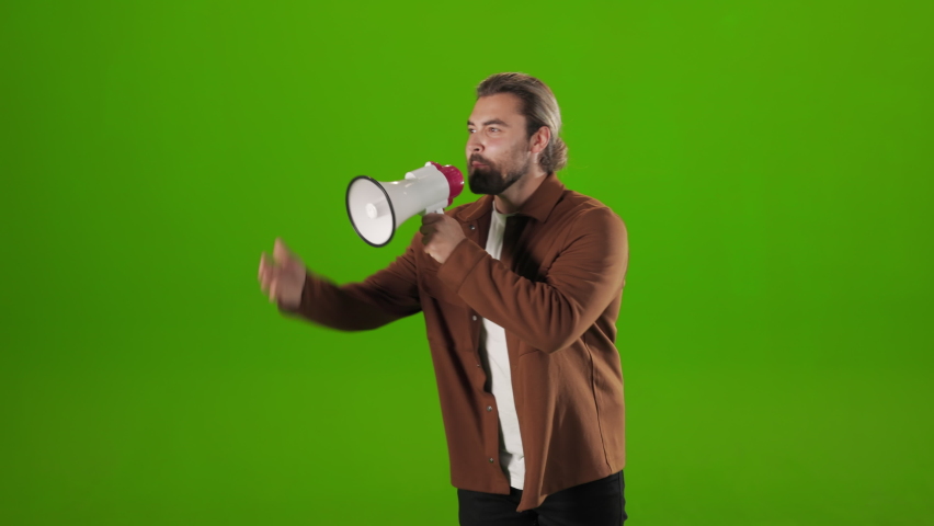 Emotional bearded man gesturing and shouting in megaphone over green background. Caucasian male hipster making loud announcement in studio.