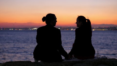 Couple on a date, young adult man and woman sitting on sea front in evening. He holds her hand and says something emotionally. Silhouettes of two people from back, red colored sunset sky in background