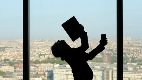 Businessman on the courage after a successful phone call, silhouette of a funny dance in front of the window. He's holding smartphone and paper folder and moving them around funny