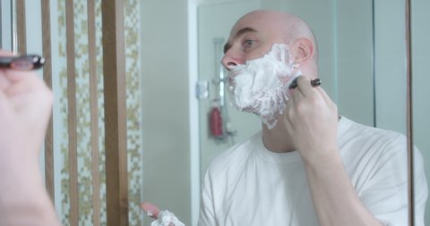 Middle-aged is shaving using disposable razor in bathroom, closeup, side view. Man applies shaving cream to face, hygienic procedure. Man is shaving beard, part of face in frame.