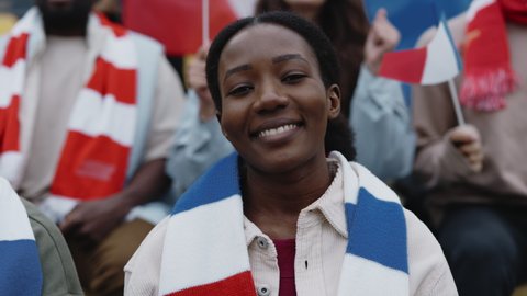 African american woman smiling and looking at camera while sitting on bleachers of stadium. Pretty lady waving with french flag while cheering during football game.