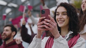 Happy female soccer fan standing on tribune with modern smartphone in hands and recording video. Young woman saving memories from live championship. - Powered by Shutterstock - Get 15% off with code: PIKWIZARD15