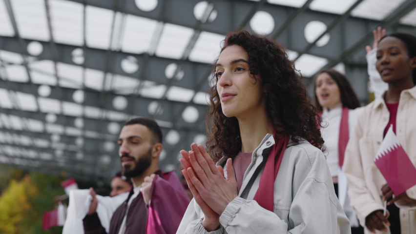Happy young woman with curly hair clapping and shouting while supporting soccer team during match on stadium. Female fan enjoying live competition among multiracial crowd on tribune.