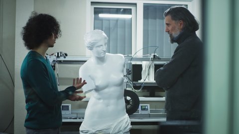 Young and mature men in casual clothes inspecting and discussing face of plastic 3D sculpture while working in modern design studio