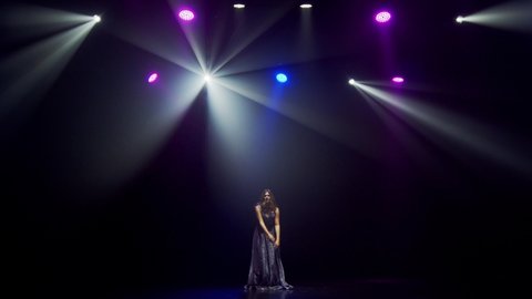 Silhouette of a luxurious singer on stage in the dark. A girl sings a song on an empty stage in the light of white, blue and purple spotlights. Bright performance and light show
