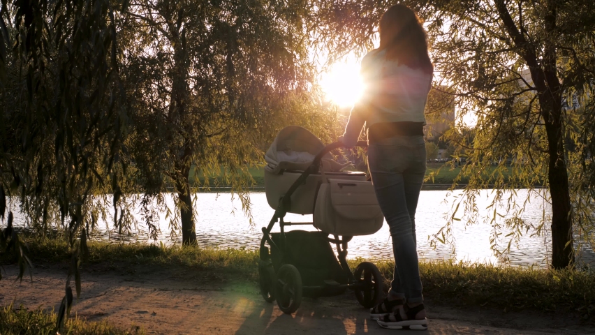 Mom babysitter on walk in park shakes stroller with baby so that he sleeps in fresh air. Background of sun at sunset, rays shine on summer evening. Relationship and care of mother for child Royalty-Free Stock Footage #1085365205