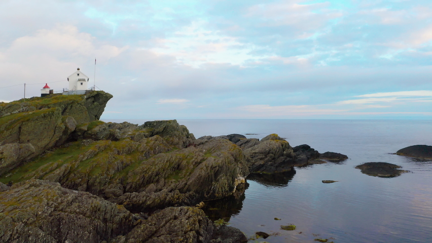 White 18th century community at oceans edge, calm circling aerial shot revealing colorful rugged shore cliffs. Bird flying away from chimney.  Endless horizon mirror the fluffy pink evening clouds.
