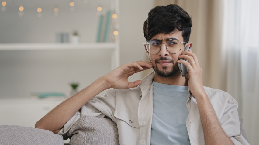 Young arab bearded man millennial guy with glasses sits on home couch talking on mobile phone answering call worries about bad connection network problems cellular trouble broken smartphone microphone Royalty-Free Stock Footage #1085374532