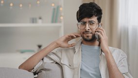 Young arab bearded man millennial guy with glasses sits on home couch talking on mobile phone answering call worries about bad connection network problems cellular trouble broken smartphone microphone - Powered by Shutterstock - Get 15% off with code: PIKWIZARD15