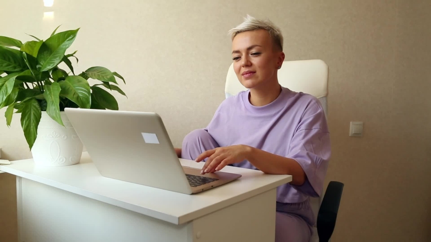a beautiful blonde girl with a short haircut is sitting at home in purple pajamas,near a laptop,typing and smiling Royalty-Free Stock Footage #1085374676