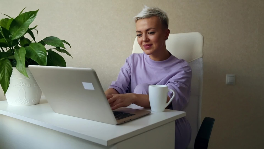a beautiful blonde girl with a short haircut is sitting at home in purple pajamas,near a laptop,typing and smiling Royalty-Free Stock Footage #1085374679