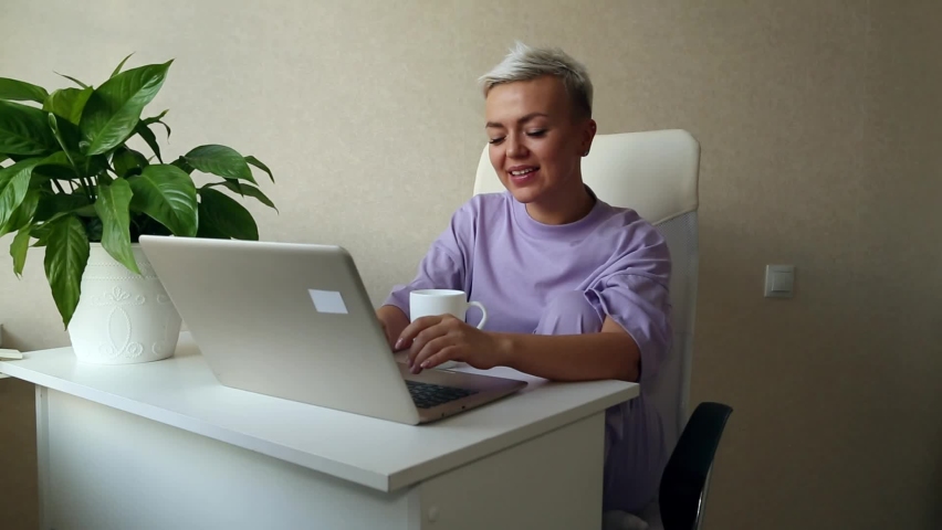 a beautiful blonde girl with a short haircut is sitting at home in purple pajamas,near a laptop,typing and smiling Royalty-Free Stock Footage #1085374682
