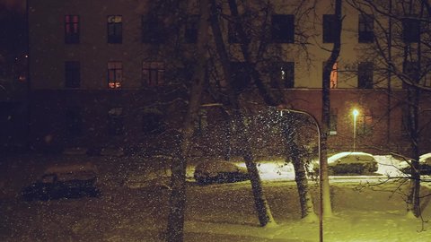 Falling snow in residential area at night. City courtyard night view in blizzard. Night town street with burning street lamps and snow covered parked cars during snowing