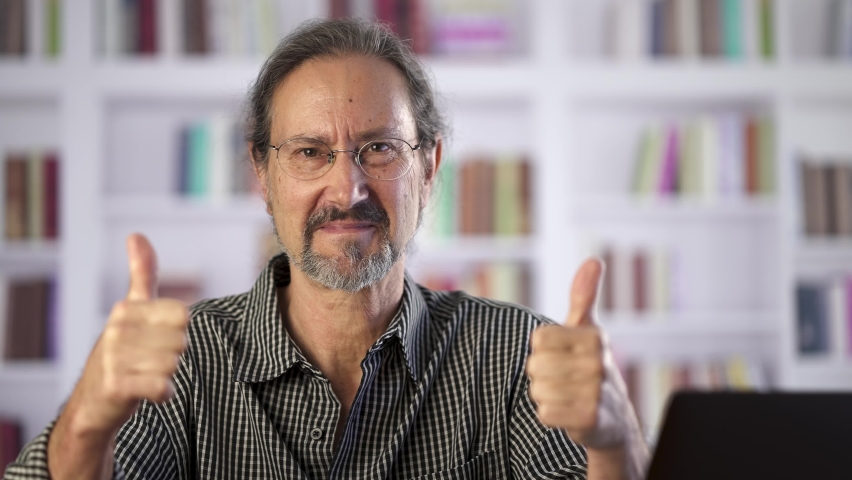 Smiling mature man gives thumbs up while at desk in library with books.