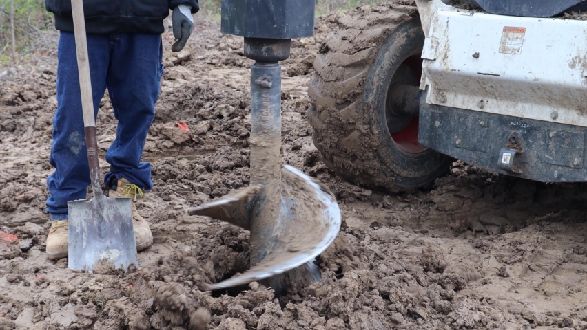 Close up of hydraulic auger mounted on skid steer loader digging a post hole for the frame of new barn construction; construction workers hold shovels; building construction, an Industrial Video by BlackBoxGuild