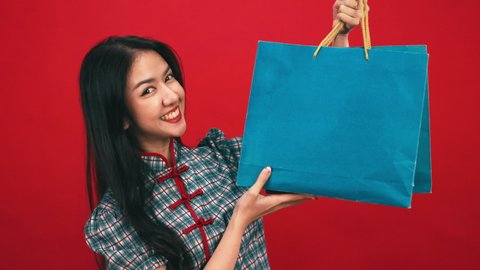 Asian woman in modern cheongsam costume holding shopping bags isolated on red studio background. Chinese new year in shopping concept style.