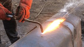 Video view of the welder through the fire. A welder in protective clothing in the open air cuts metal pipes with acetylene welding and fire and sparks are visible.