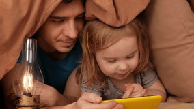 Father and daughter watch cartoon on phone, home interior, training. Smartphone with internet, funny video. Modern technology, bedroom. lie in house