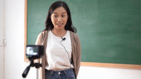 Smiling Asian female teacher live teaching online with video camera in classroom. She recording video tutorials for her video blog for students. Online education.