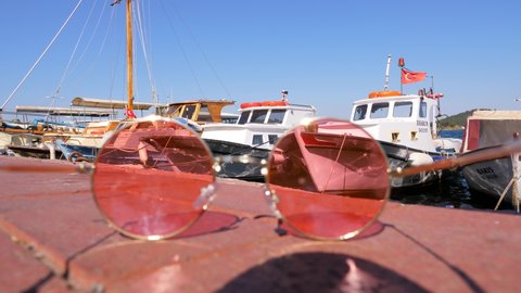 ISTANBUL - AUGUST 16, 2021: Fishing boats rocking on water by pier, view through rose-colored glasses lying on pavement. Focus in background, glasses out of focus. Conceptual shot of Burgazada