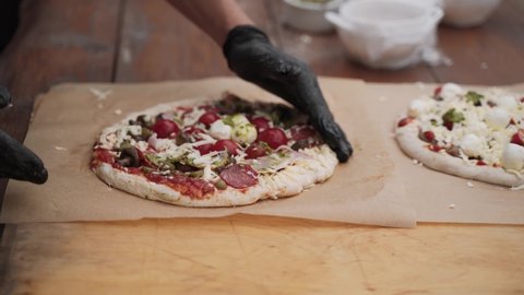 Unrecognizable man in black gloves, pizzaiolo fix raw pizza dough and points to filling. Appetizing dish with ingredients mozzarella, tomato, sauce, pepperoni lies on baking paper, parchment.
