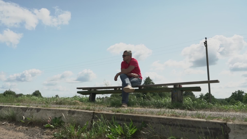 adult male sits on bench of bus stop in rural area waiting for bus and looks at watch holding phone