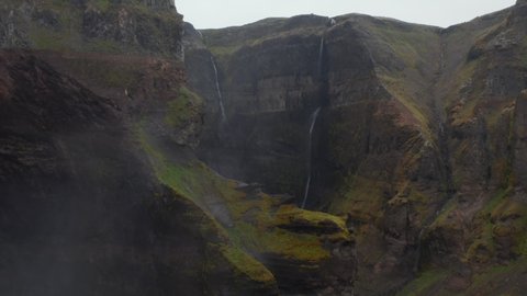 Deep rocky canyon with tall steep sides overgrown by green vegetation. Backwards reveal of tall waterfall. Beautiful natural scene. Haifoss, Iceland
