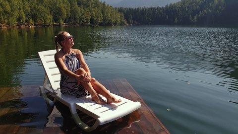 Woman lie on a sunbed in sunglasses and a boho silk shawl. Girl rest on a flood wood underwater pier. The pavement is covered with water in the lake.