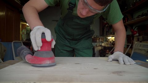 Carpenter grinds wooden board with orbital sander machine at workshop